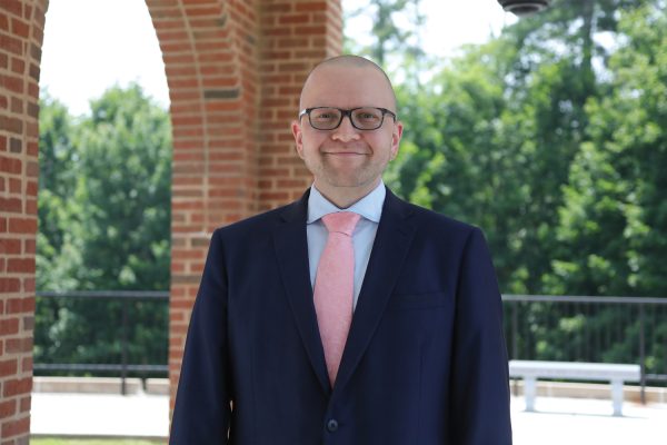 A picture of Brandon Gilbert, Creative Services Manager, wearing a dark blue suit, light blue shirt, and pink tie, while standing outdoors at University Point.