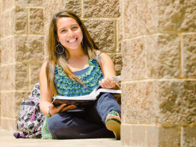 Student sitting against a brick wall smiling and reading a textbook