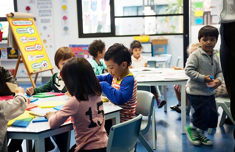 A group of diverse students sitting at tables in a daycare playing with colorful shapes and reading picture books