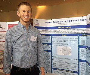 Andrew Thomas standing next to his poster presentation for his research titled College: Casual Sex or Old School Dating