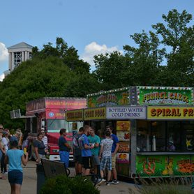 A photo of several people in front of food trucks with Concord University's Marsh Hall bell tower in the background behind some trees