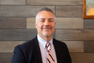 A photo of Andrew Sulgit, Government Relations & Major Gifts Officer at Concord University. Andrew is standing front of a gray slate tile wall and is wearing a dark blue suit with a pink and blue-striped tie.