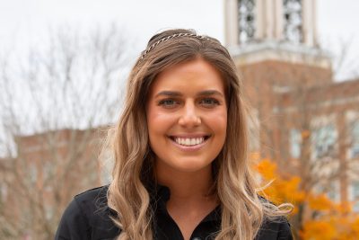 A photo of Lilly Fleming, a Concord University Admissions Counselor, with an orange Fall foliage backdrop, with the iconic Concord bell tower in the background, against a dark blue moody October sky.