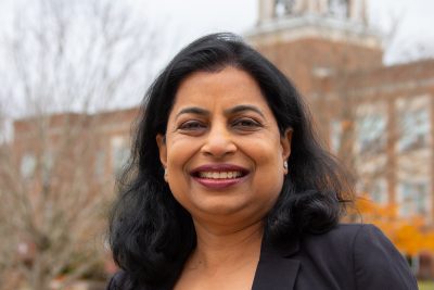 A photo of Vartika Jain, with an orange Fall foliage backdrop, with the iconic Concord bell tower in the background, against a dark blue moody October sky.
