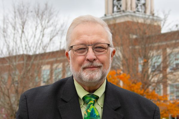 A photo of James (Jim) Owston, with an orange Fall foliage backdrop, with the iconic Concord bell tower in the background, against a dark blue moody October sky.