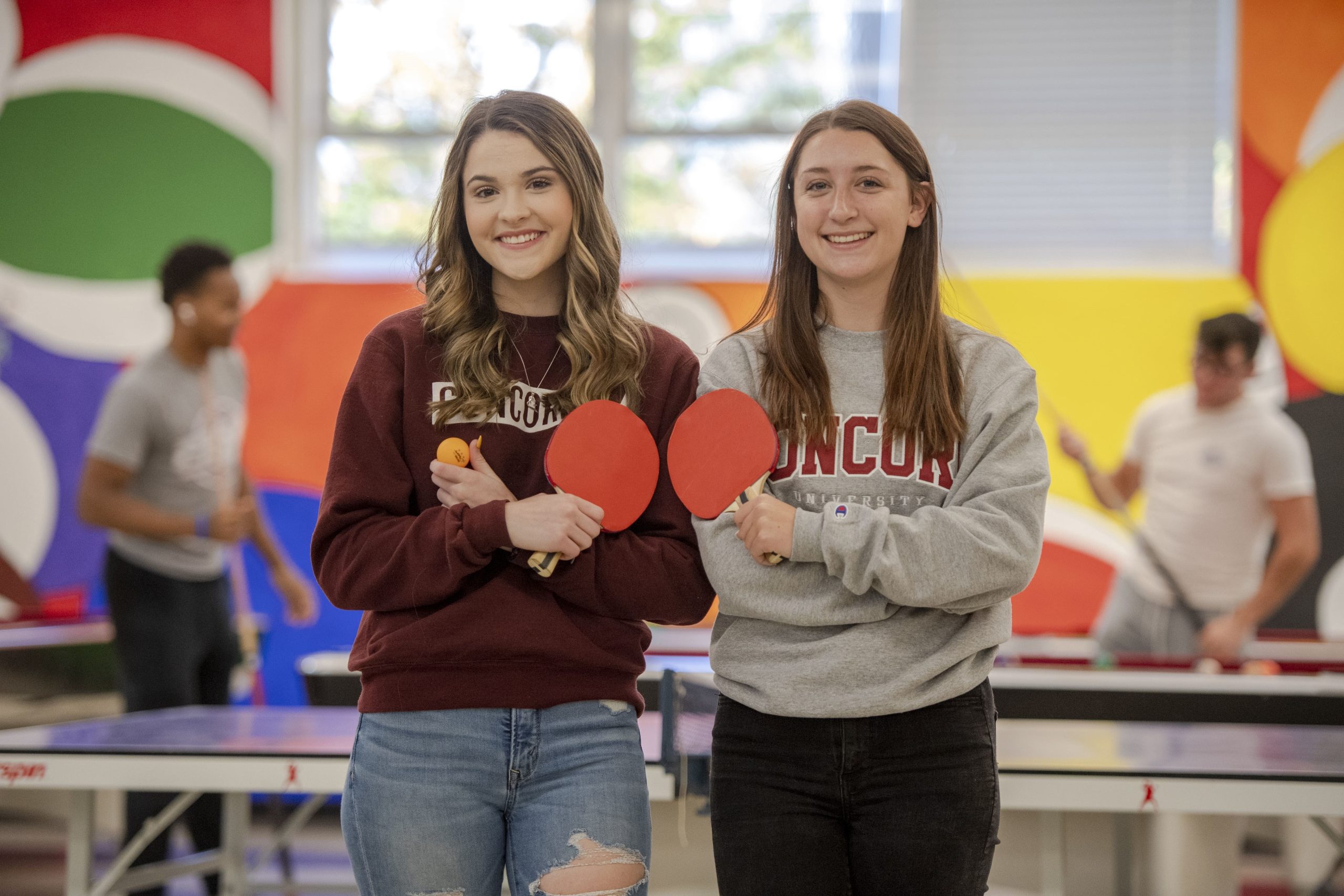 Two students holding table tennis paddles in Concord University's game room. There are two additional students playing pool in the background