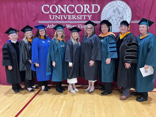 Administration, nursing faculty, and BSN graduates pose together in front of a grand Concord University ceremonial curtain.